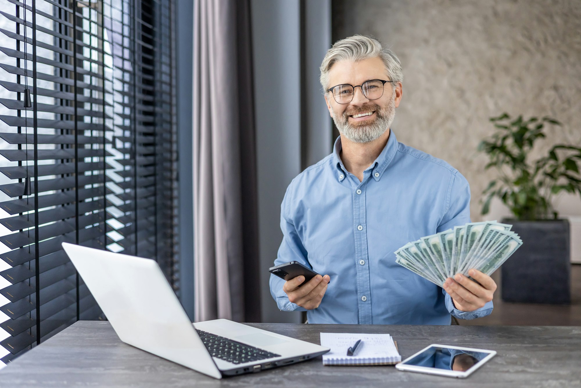 Successful mature entrepreneur with cash and smartphone in modern office, financial success concept